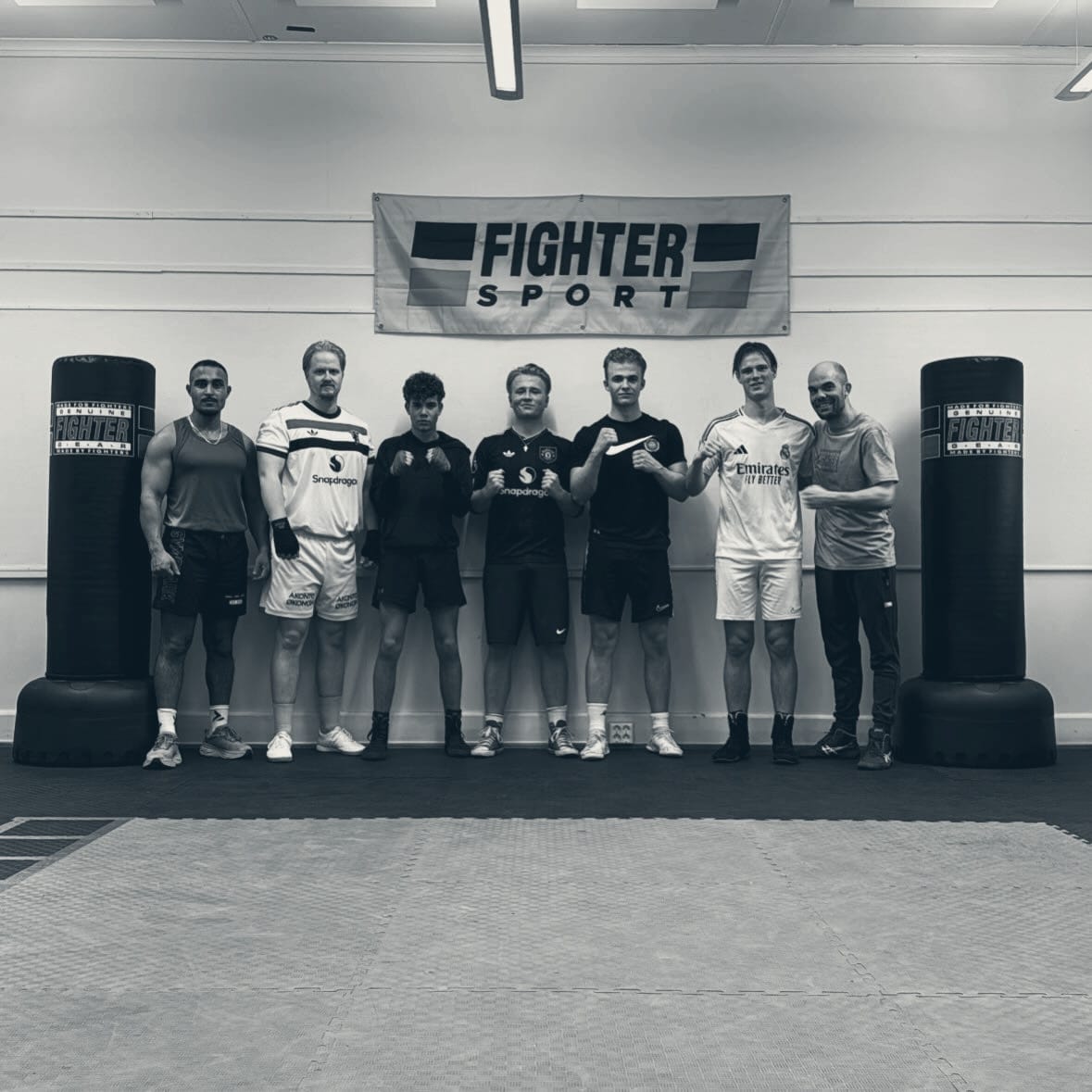 Sju boksere fra Omie's Gym på rad foran Fighter Sport-banneret, mellom to frittstående sekker i klubblokalet i Hønefoss